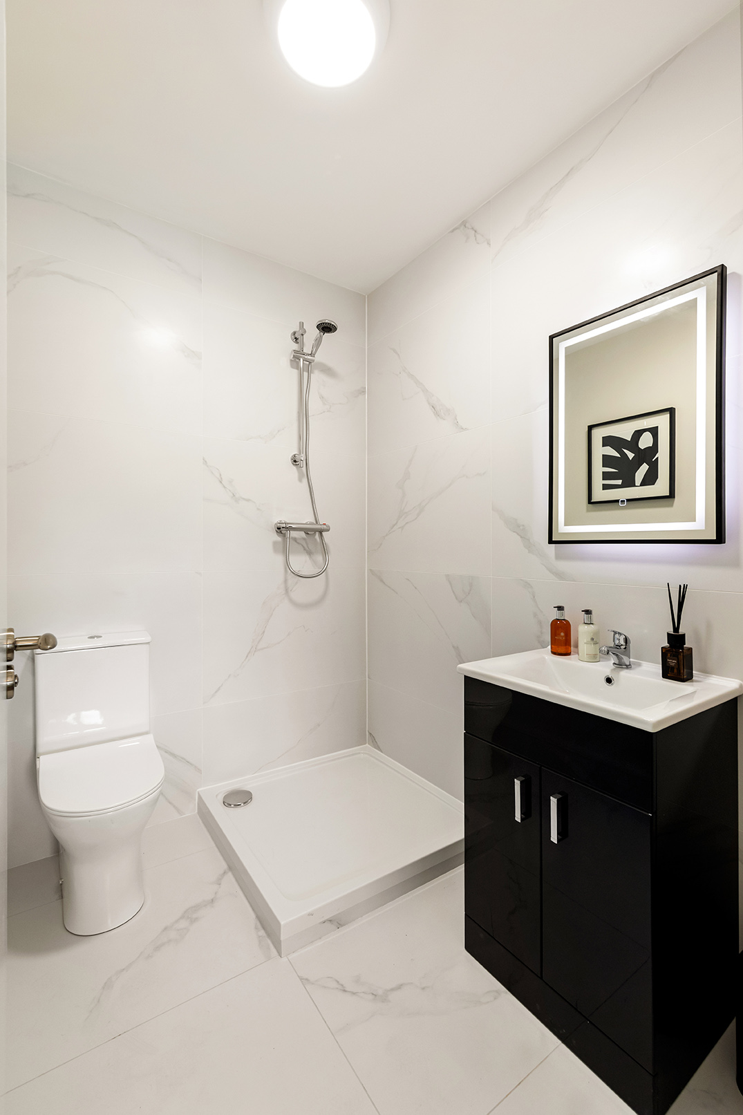 Modern bathroom with white marble walls, a shower area, toilet, black vanity with sink, and a backlit mirror.