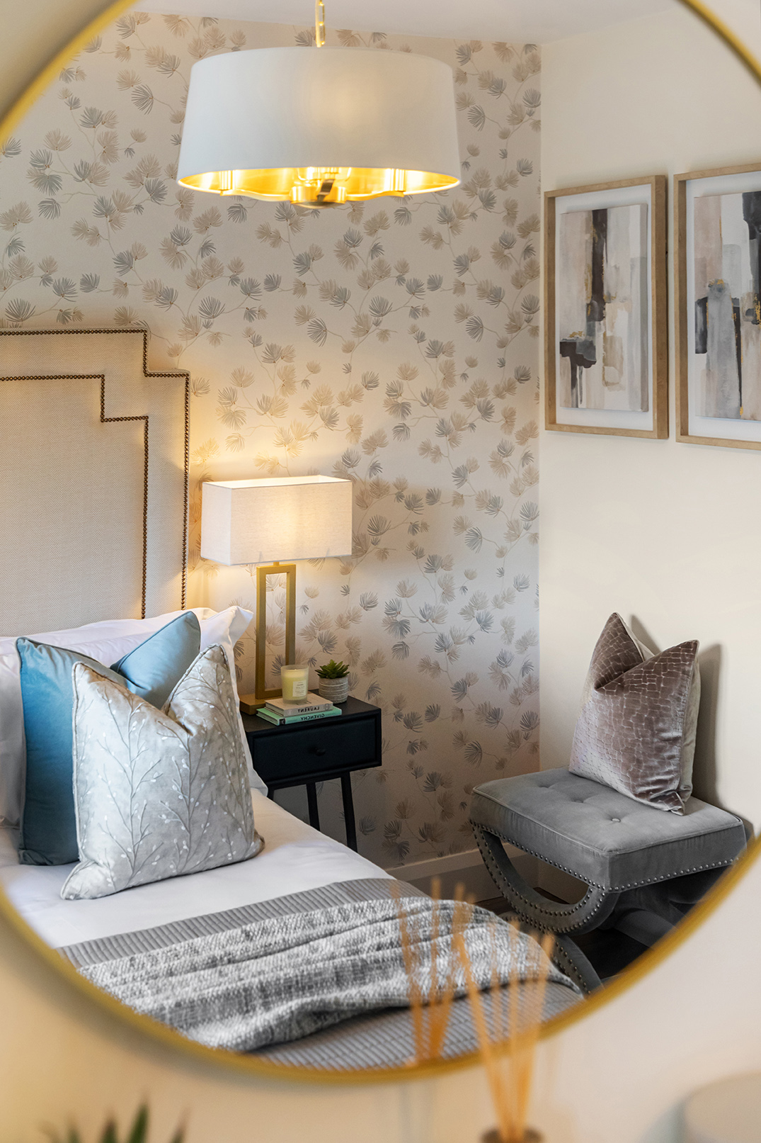 Reflection of a bedroom corner with patterned wallpaper, a bed with blue and beige pillows, a bedside table with a lamp and books, and a gray upholstered bench with a velvet pillow.